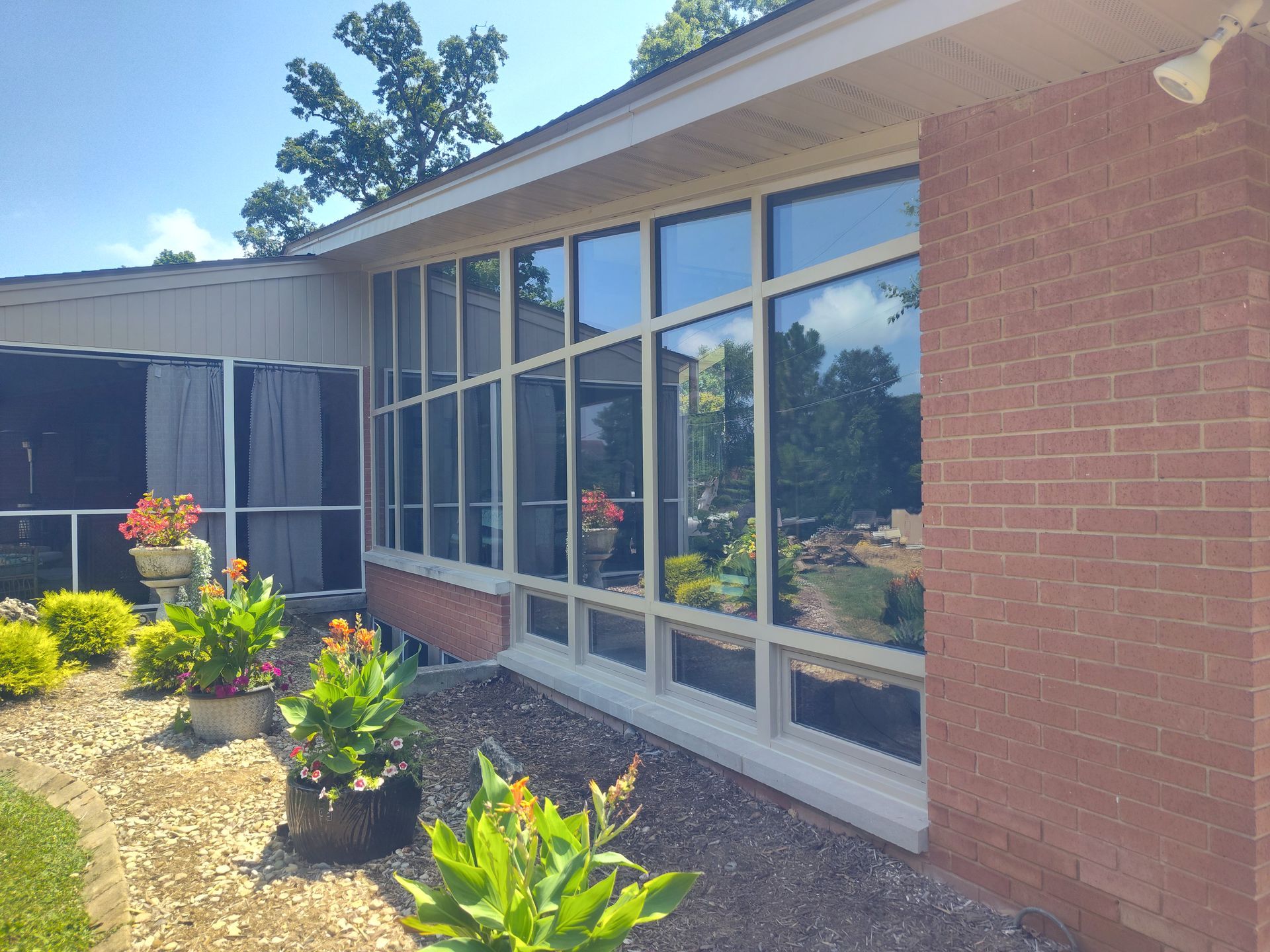 A sunroom with many windows reflecting trees and sky. Red brick wall and bushes with flowers.