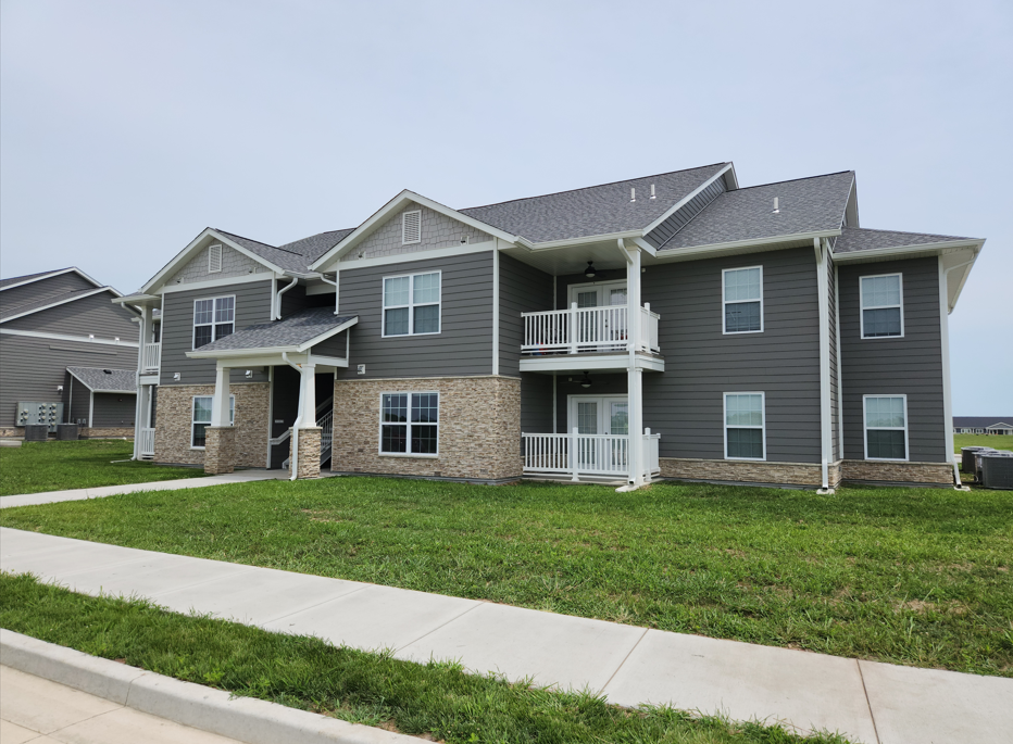 Apartment building with grey siding, brick base, white trim, balconies, and green lawn under a cloudy sky.