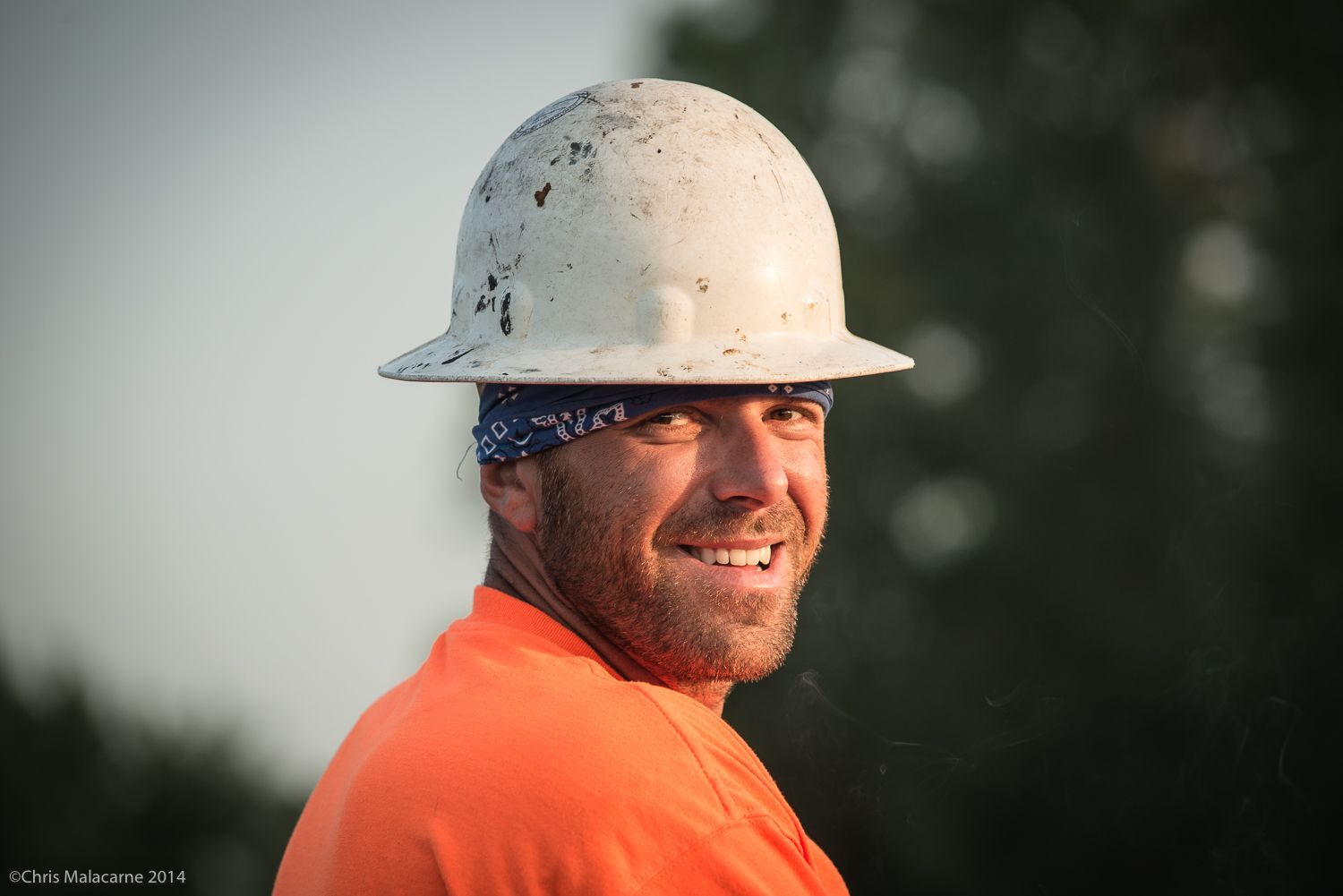 A man wearing a hard hat and an orange shirt is smiling.
