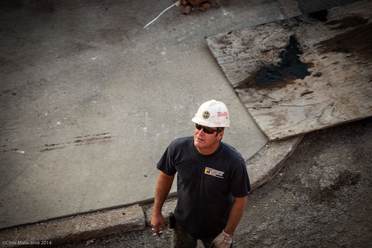 A man wearing a hard hat and sunglasses is standing on a sidewalk.