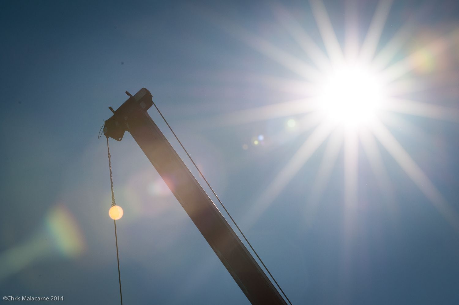 A crane is silhouetted against a blue sky with the sun shining brightly behind it.