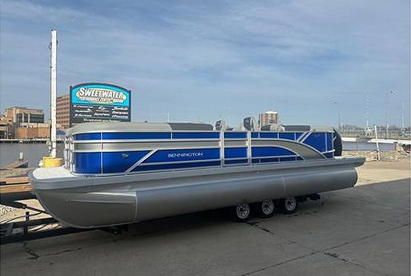 Blue and silver pontoon boat on a trailer, sunny day. The boat is near a body of water, and a sign is visible in the background.