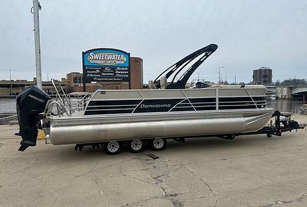 Pontoon boat on trailer, near a building and water. Silver and tan, with black motor.