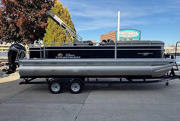 Pontoon boat on trailer, silver and black, with a motor, parked outside near building.