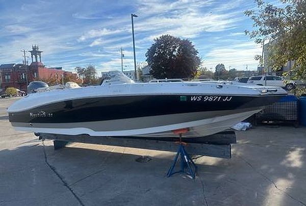 White and black motorboat on a trailer, parked on a concrete surface with buildings and blue sky in the background.