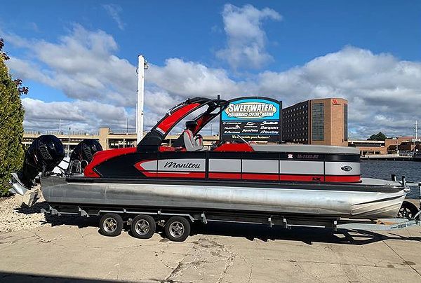 A red and black pontoon boat on a trailer, next to a waterfront with a city backdrop.