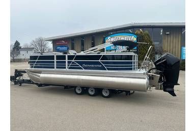 A large pontoon boat on a trailer parked outside a building. The boat is silver and black with a black outboard motor.