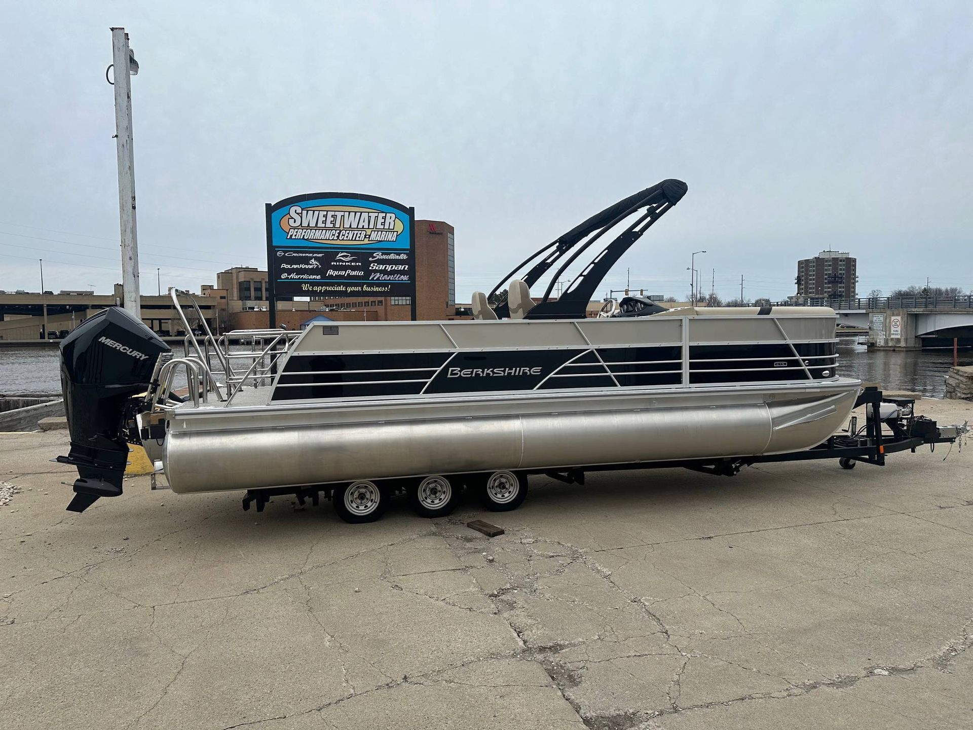 Pontoon boat on trailer, beige and black, with outboard motor, parked near water.