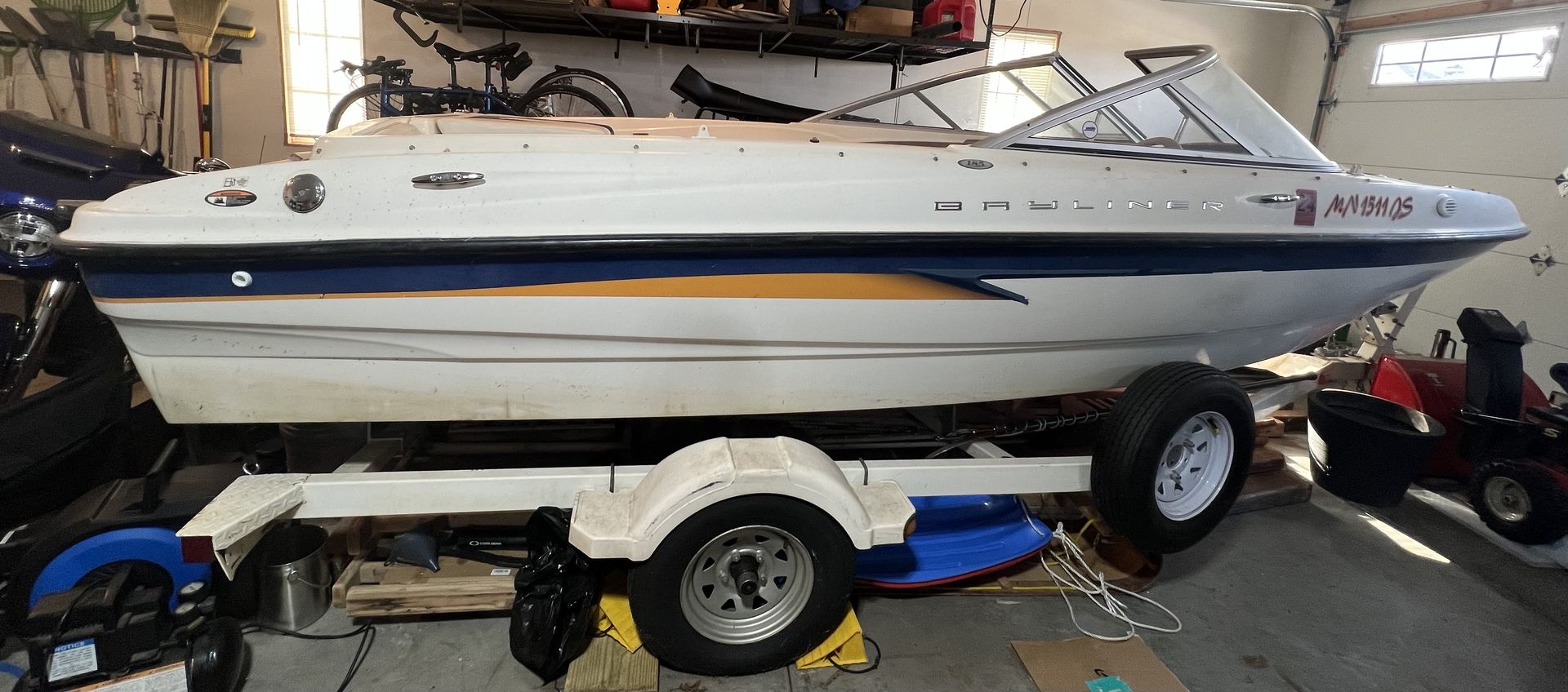 Boat on a trailer in a garage. White boat with blue and yellow accents. Red lawn mower to the right.