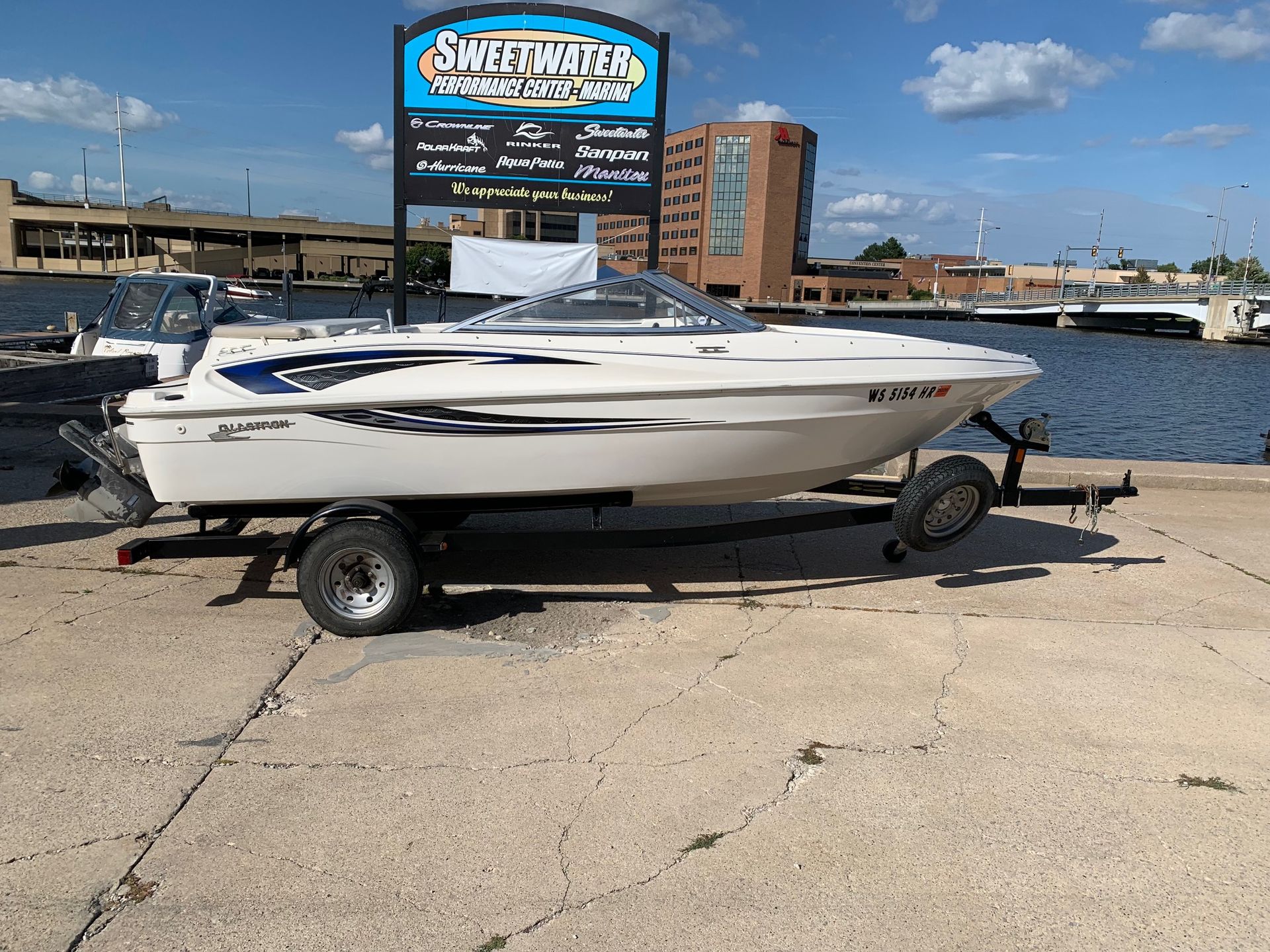 White boat on a trailer in front of a Sweetwater sign, water, and buildings.