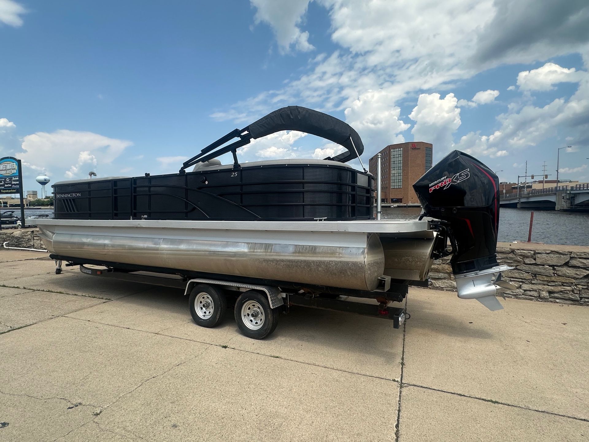 Pontoon boat on trailer, black canopy, large motor, parked on concrete near water.
