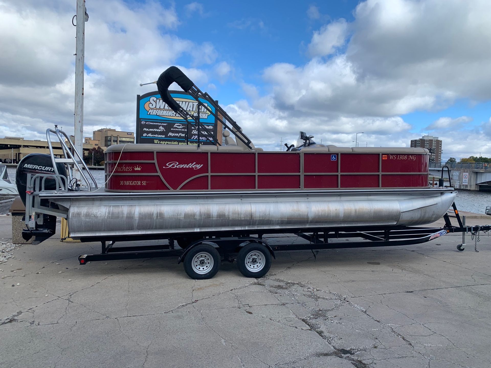 Red pontoon boat on a trailer, parked on concrete near a body of water and a building.