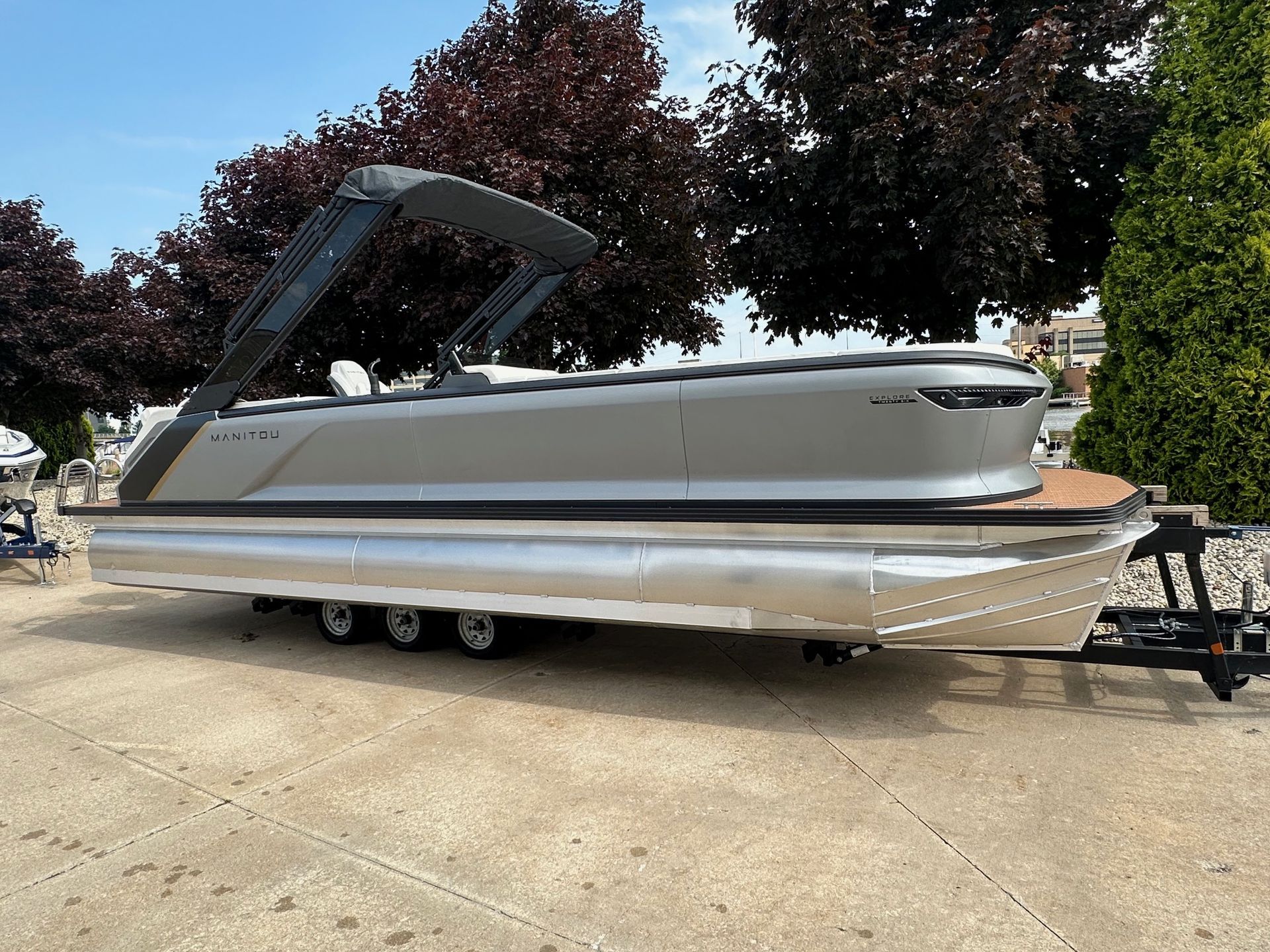 Silver pontoon boat on a trailer, with a gray canopy, parked outside on a sunny day.