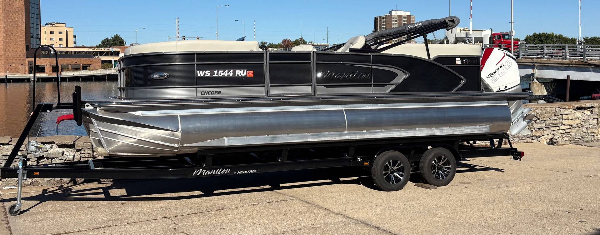 A black pontoon boat on a trailer, parked on a gravel lot near a body of water.