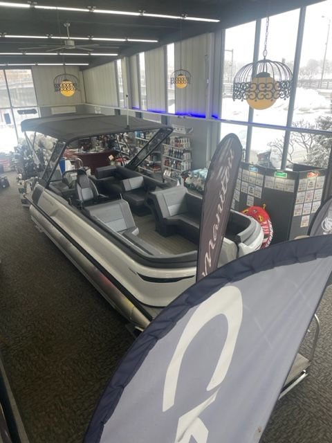 A white and gray pontoon boat on display inside a showroom, with a partially visible blue sign.
