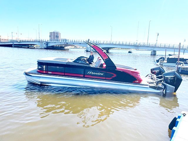 Pontoon boat with red accents floating in water near a bridge.