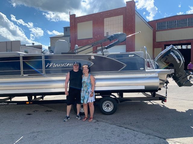 A couple stands in front of a new pontoon boat on a trailer, outside a brick building on a sunny day.