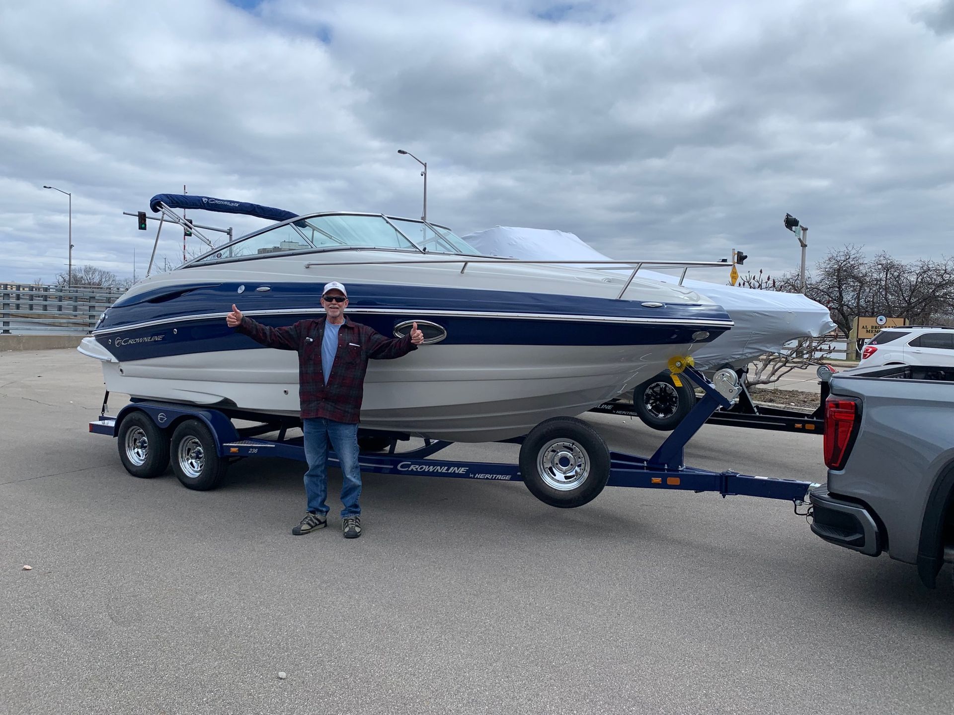 Man stands proudly next to a boat on a trailer, arms outstretched. Cloudy day, gray asphalt. Blue and white boat.