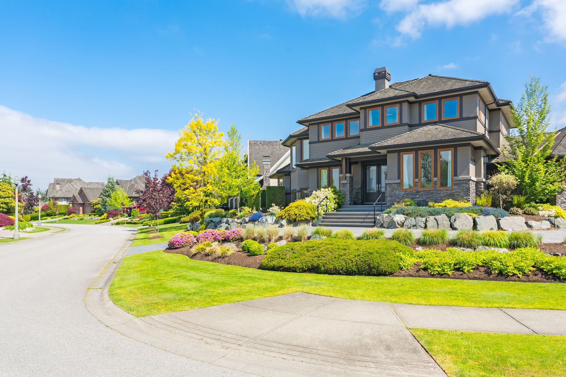 A two-story grey suburban house with stone accents, a landscaped front lawn, and trees under a clear blue sky.