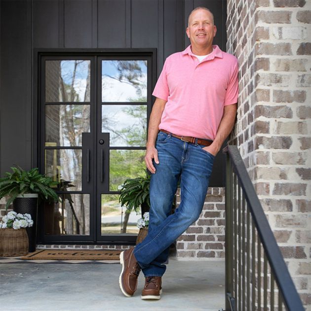 Man in a pink polo and jeans leaning by a brick entryway with black doors and potted plants