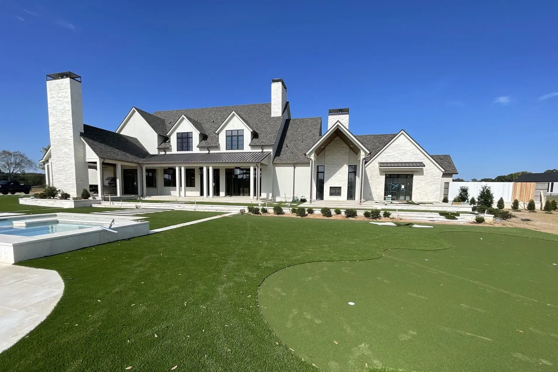 Large white home with a dark shingled roof, tall chimneys, and a backyard putting green under a bright blue sky.