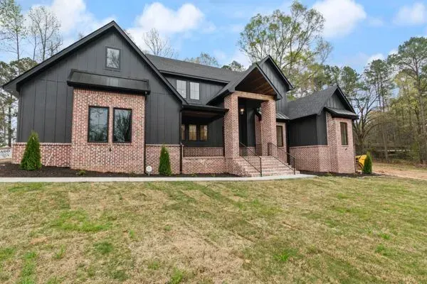 A two-story house with dark siding and brick accents, featuring a front porch, a gabled roof, and a grassy yard.