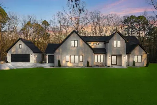 A large, modern, two-story house with a light-colored stucco exterior, black roof, and a lawn under a sunset sky.