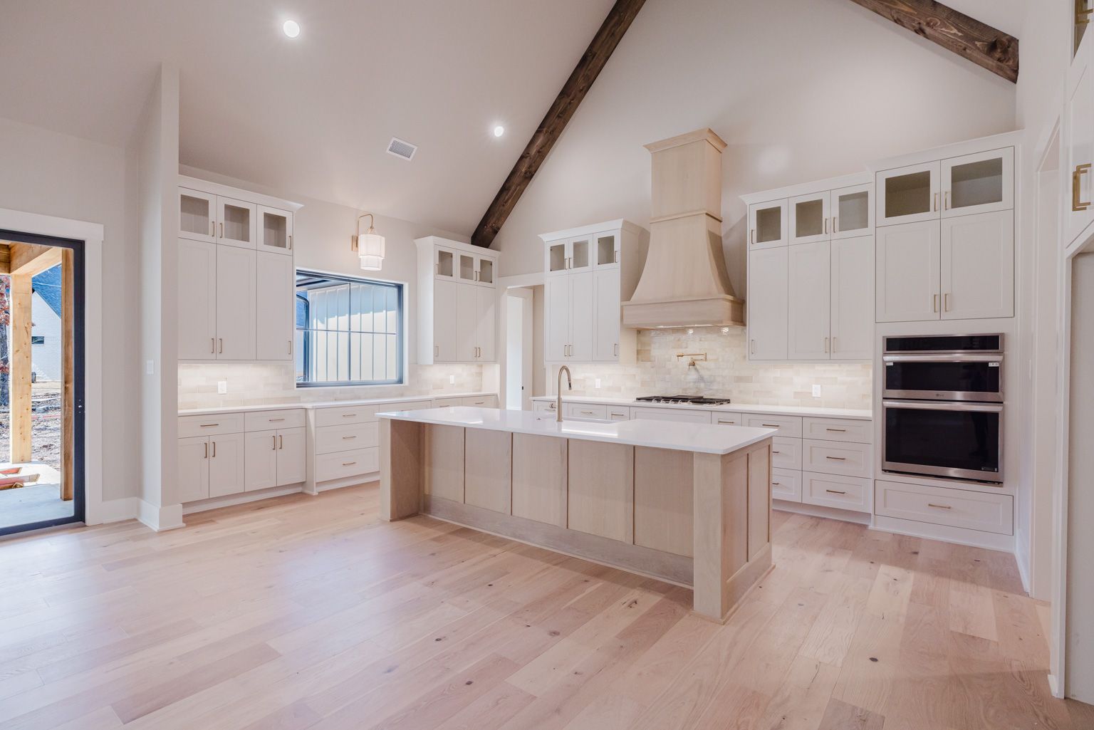 A bright, modern kitchen with white cabinets, a light wood kitchen island, stainless steel double ovens, and wood beams.