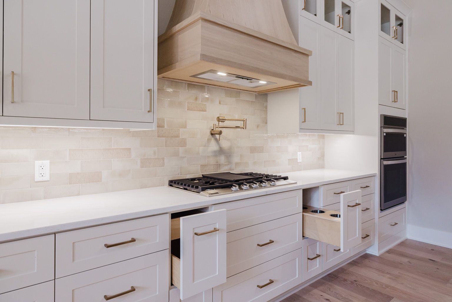 A modern kitchen featuring white cabinets, a light wood vent hood, a neutral tiled backsplash, and open pull-out drawers.