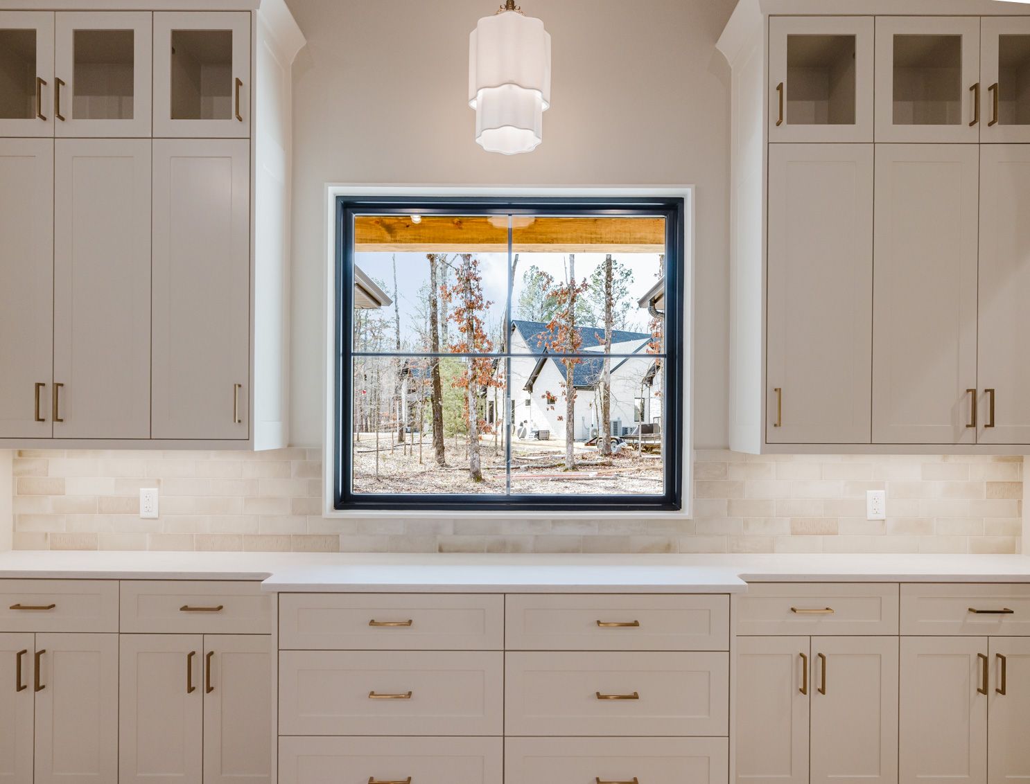 Light-colored kitchen cabinets flank a central black-framed window with a pendant light hanging above, set over a countertop.