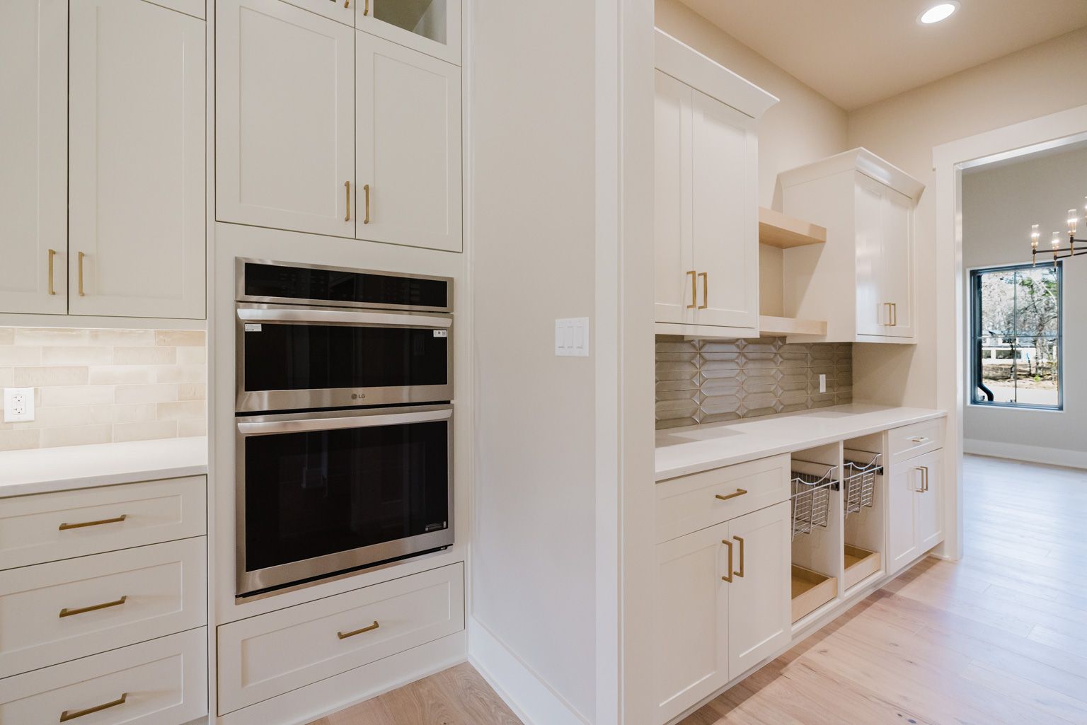 A bright, modern kitchen features white cabinetry, a double wall oven, and light wood flooring leading to another room.