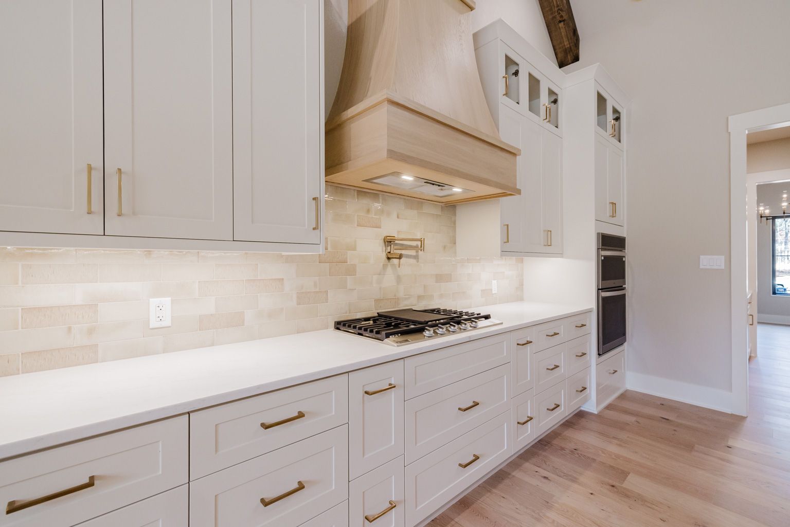 A bright kitchen featuring white shaker cabinets, a light wood range hood, a beige stone backsplash, and quartz counters.