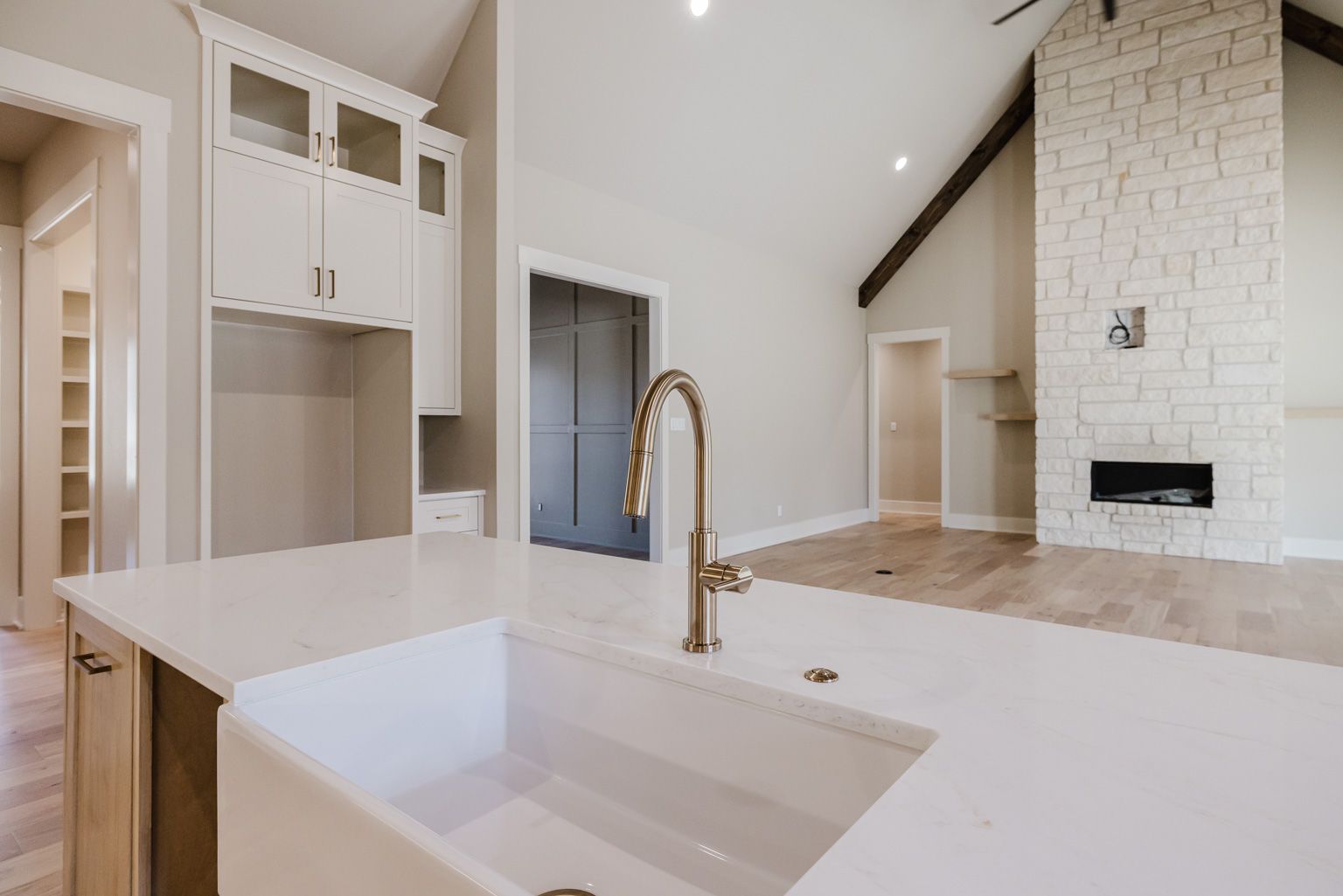 A white farmhouse kitchen island with a gold faucet, overlooking a bright open living area with a stone fireplace.