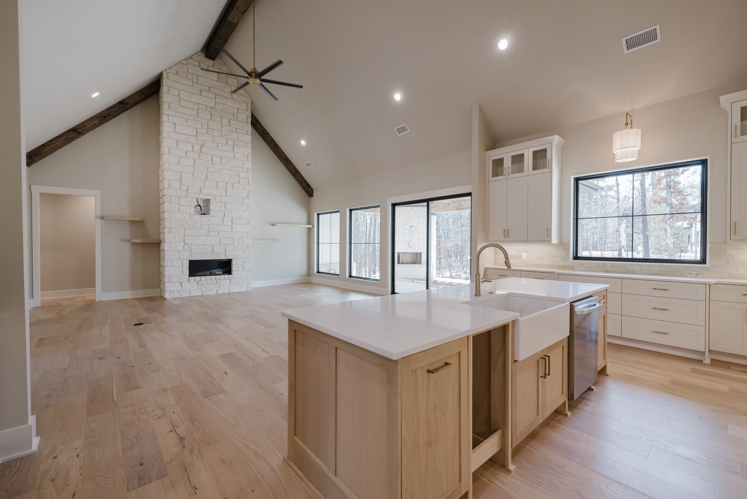 A bright open-concept kitchen and living area featuring a stone fireplace, light wood floors, and a kitchen island.