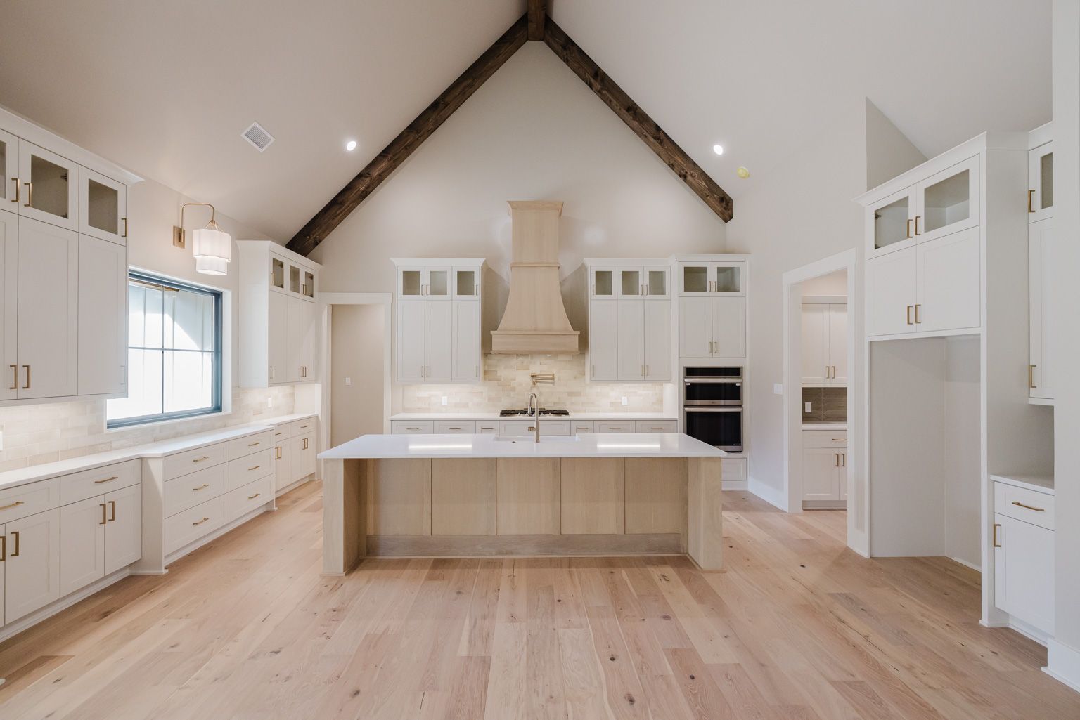 A modern, high-ceiling kitchen with white cabinets, a central island, light wood floors, and exposed wooden ceiling beams.