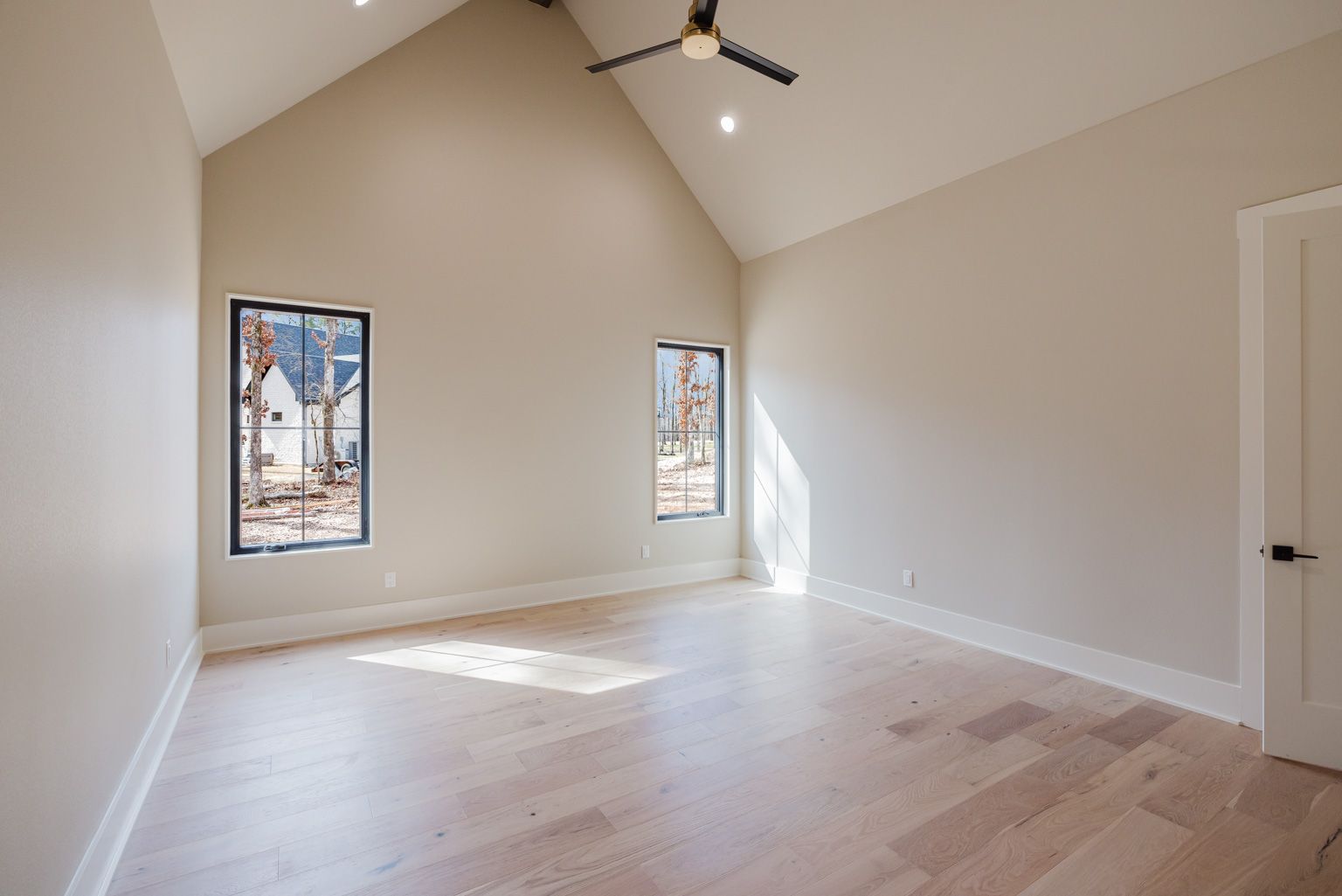 A bright, empty room with light wood floors, beige walls, a vaulted ceiling, a ceiling fan, and two windows.