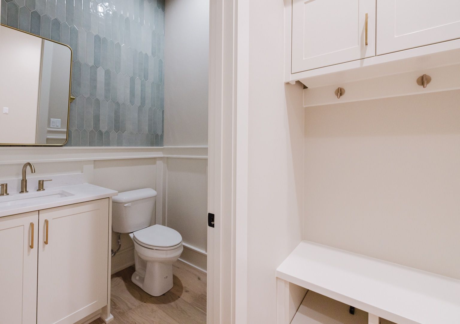 A bathroom with a white vanity, a toilet, a blue-tiled wall, and an adjacent entryway with a bench and overhead cabinets.