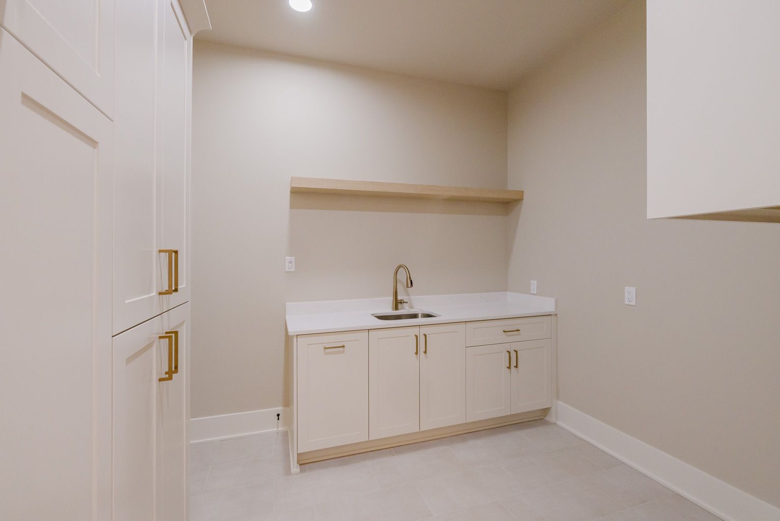 A laundry room with white cabinets, gold hardware, a light stone countertop, a sink, and a floating wood shelf.