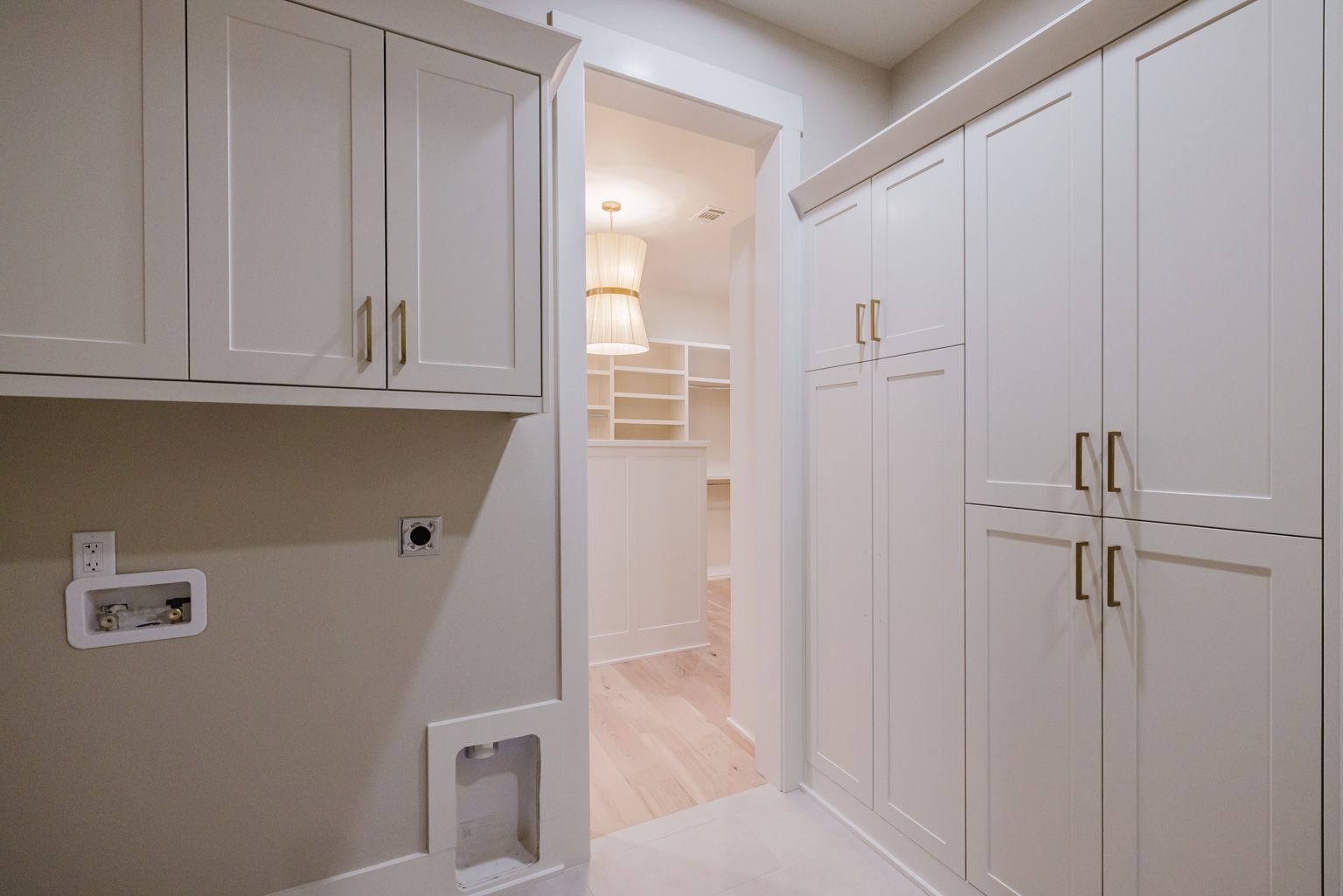 A modern laundry room with white cabinetry and a door opening into an adjoining room with light wood floors.