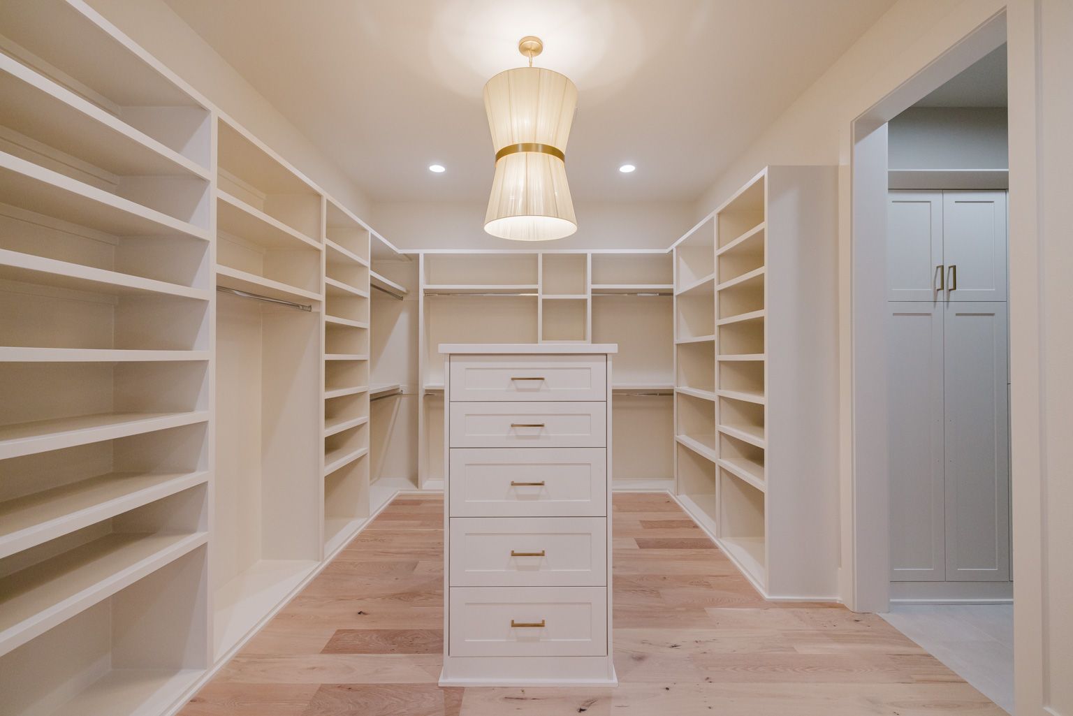 A walk-in closet with white shelving, a central five-drawer dresser, light wood flooring, and a tiered pendant light.