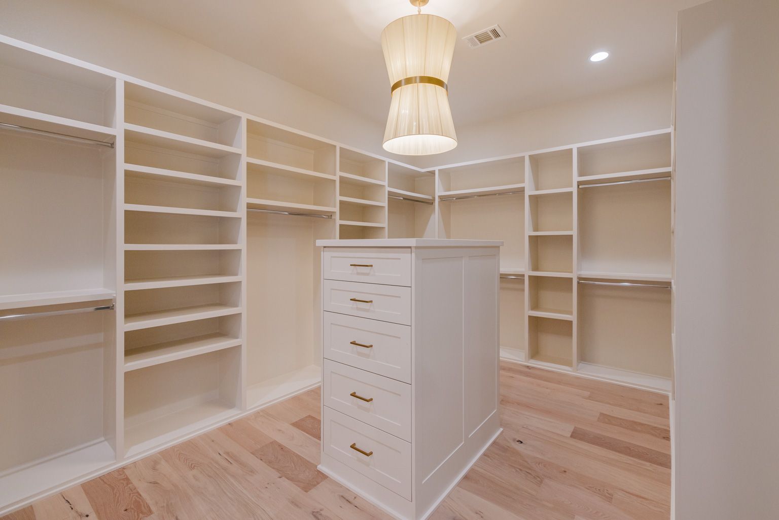 A walk-in closet featuring white shelves, hanging rods, a central dresser island, and a pendant light above.