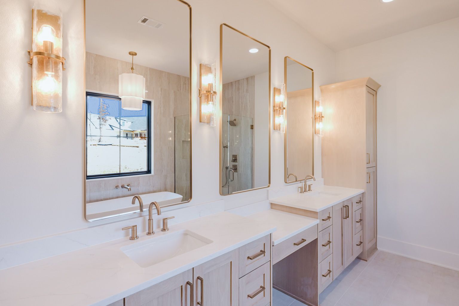 A bright, modern bathroom featuring light wood cabinets, white countertops, three tall mirrors, and wall-mounted lights.