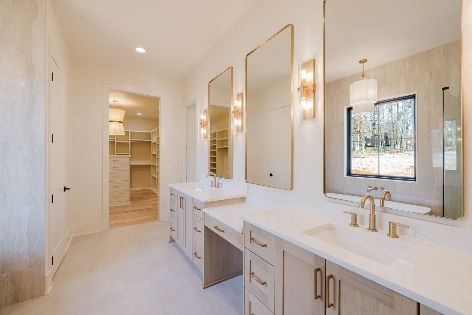 A bright, modern bathroom featuring a double vanity with light wood cabinets, gold hardware, and large wall-mounted mirrors.