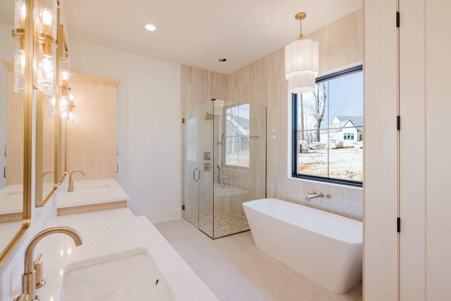 Modern bathroom with a double vanity, gold fixtures, glass-enclosed shower, freestanding tub, and a window.