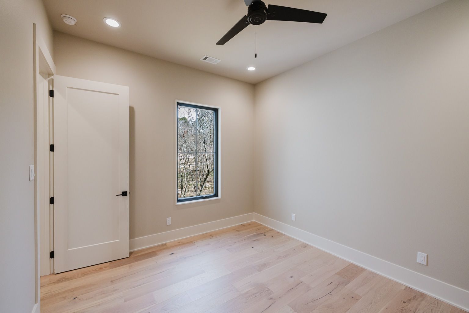 An empty room with light wood floors, beige walls, a single rectangular window, and a dark ceiling fan.
