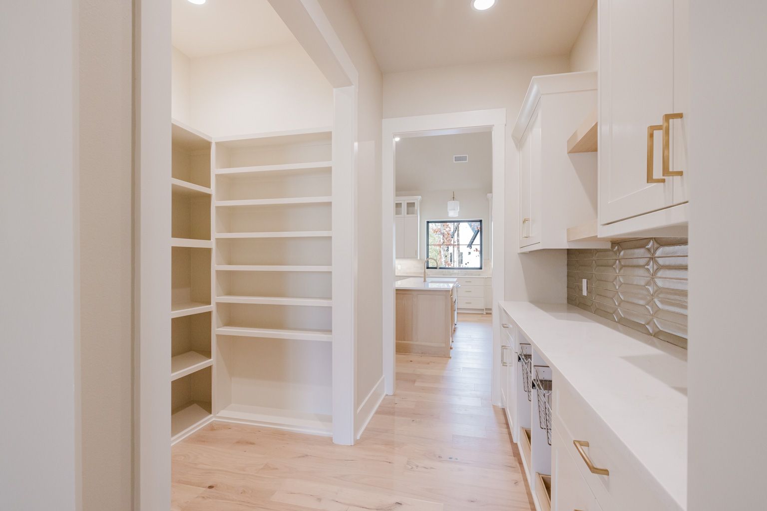 A bright pantry with white shelving, a long counter with a tiled backsplash, and an opening to a kitchen beyond.