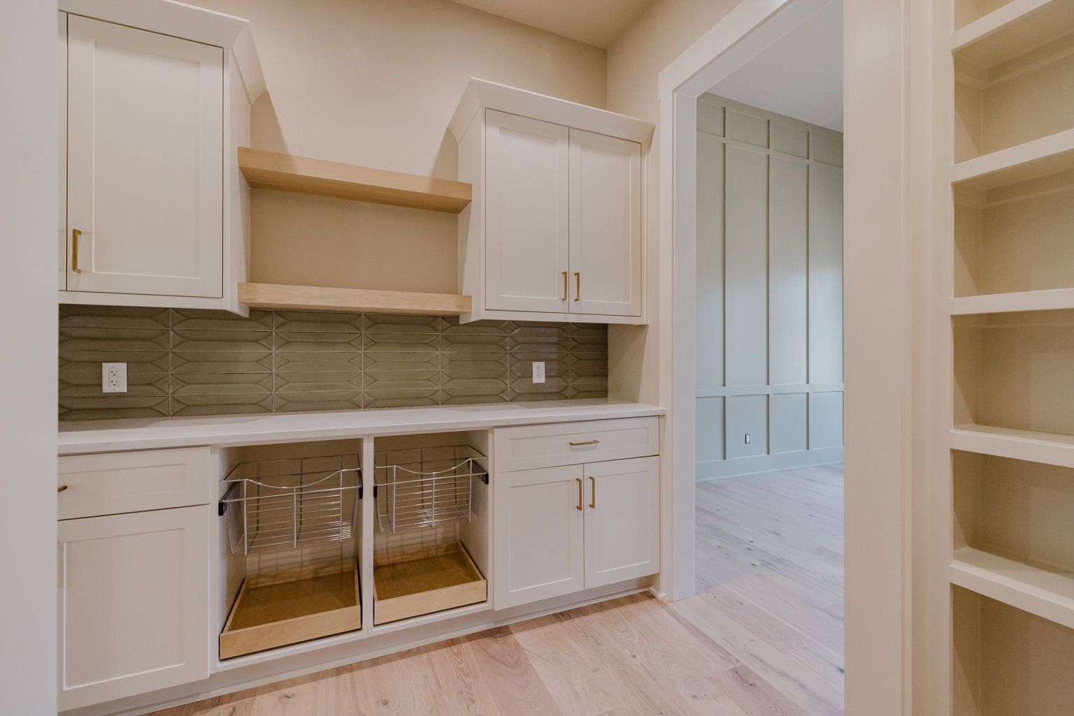 A kitchen butler’s pantry with white cabinets, wood-tone open shelving, gray tile backsplash, and built-in wall shelving.