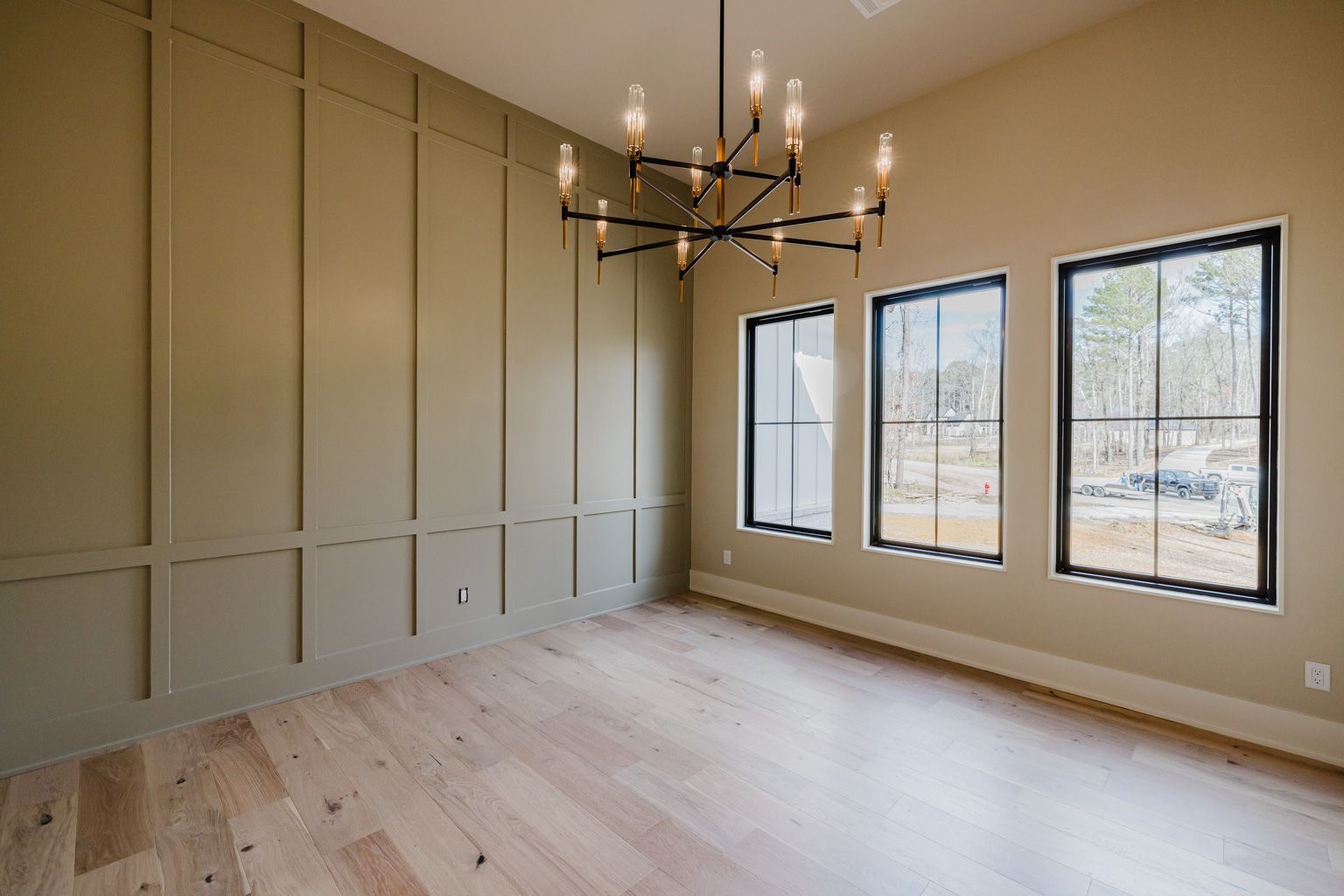 A modern room with olive green board-and-batten accent wall, light wood floors, three black-framed windows, and chandelier.