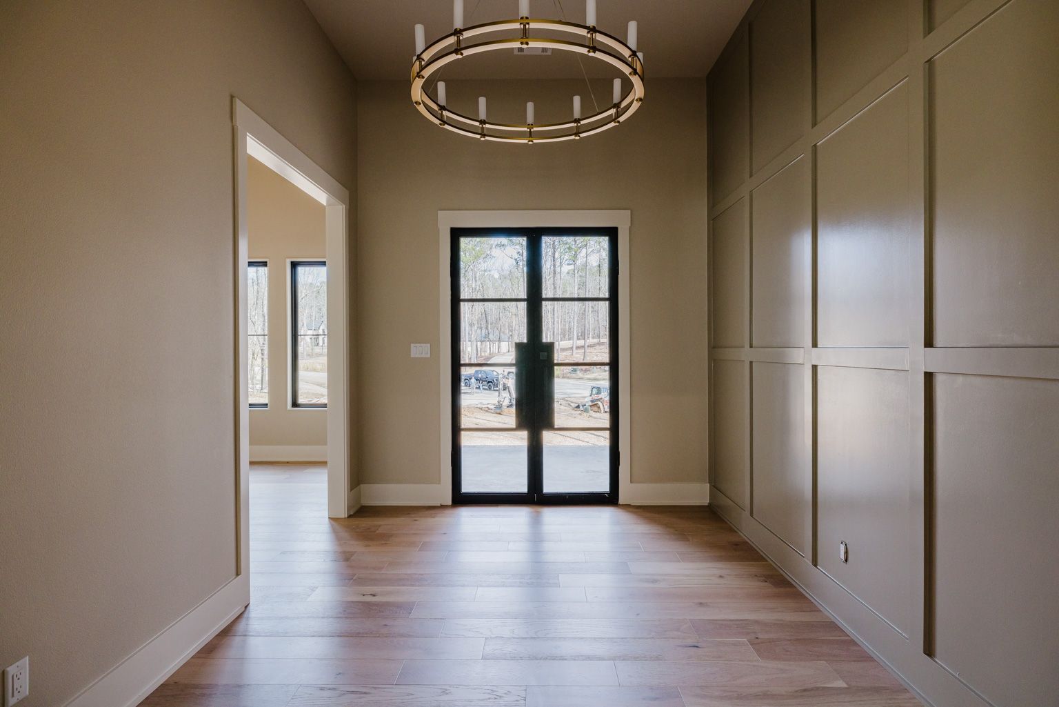 A foyer with light wood floors, a gold wheel chandelier, a paneled wall, and glass double doors leading to a bright exterior.
