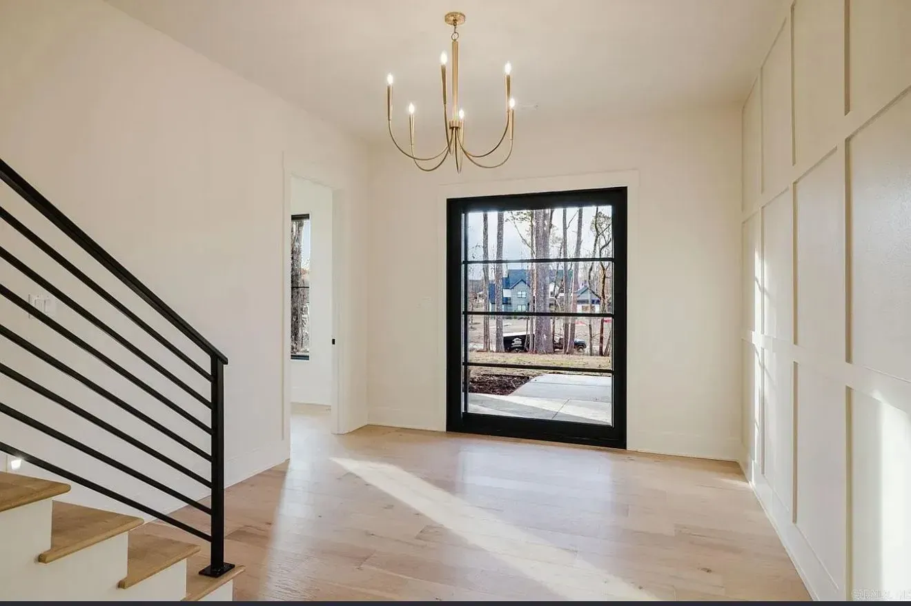 A bright entryway featuring light wood floors, a modern black chandelier, a staircase with a black railing, and a window.